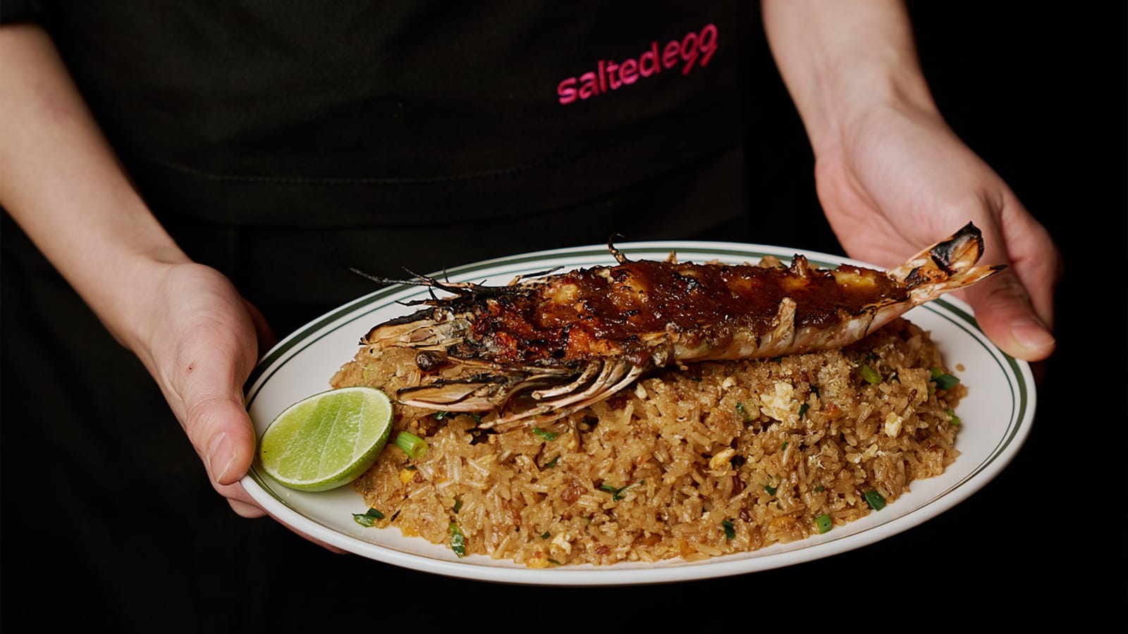 A whole grilled king prawn over fried rice with lime, held by a Salted Egg chef